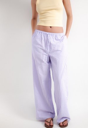 Personne portant un pantalon blanc ample avec de fines rayures verticales, un top court jaune sans manches et des sandales marron, debout les mains dans les poches.