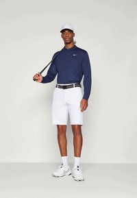 Mann med hvit caps, marineblå langermet polo, hvite shorts og joggesko, som holder en golfkølle over skulderen, står mot en ensfarget bakgrunn.