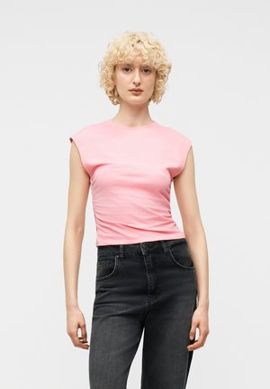 Blondynka o kręconych włosach, ubrana w dopasowany różowy top bez rękawów i czarne jeansy z wysokim stanem, stojąca na jednolitym tle.