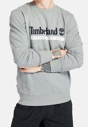 Osoba nosząca szarą bluzę Timberland z skrzyżowanymi, wytatuowanymi ramionami i czarnymi spodniami, docięta przy szyi i talii.
