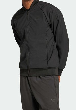 Homme portant une veste de survêtement Adidas noire et un pantalon assorti avec un logo discret et des poches zippées, debout devant un fond clair uni.