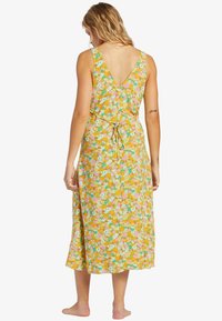 Robe d'été fleurie avec un dos en V, fabriquée en tissu léger. Présente un fond jaune avec des fleurs colorées et un détail de taille à nouer.