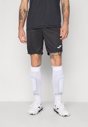 Sorte sports shorts med hvide sidelogoer, parret med hvide fodboldsokker med teksturmønster og sorte fodboldstøvler.