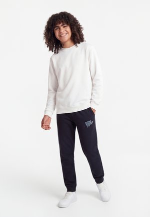 Lachende jongere met krullend haar, gekleed in een witte trui, zwarte joggingbroek met afbeelding en witte sneakers, staand op een witte achtergrond.