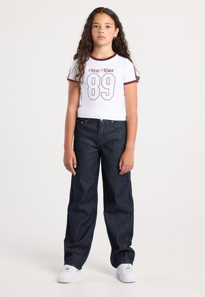 Meisje met lang krullend haar, gekleed in een wit "New York 89" t-shirt, donkere streepjessenbroeken en witte sneakers, staand tegen een eenvoudige achtergrond.