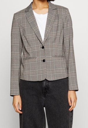 Woman wearing a gray plaid blazer with two black buttons over a white shirt and black pants, standing against a plain background.