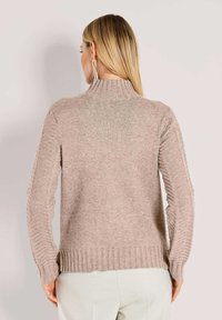 Frau mit blonden Haaren trägt einen beigen Strickpullover mit geripptem Kragen und Bündchen sowie cremefarbenen Hosen und steht vor einem hellen Hintergrund, Rückansicht.