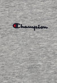Tessuto di cotone grigio con il logo ricamato "Champion" in blu navy con un accento rosso. La texture è liscia e leggermente melangiata.