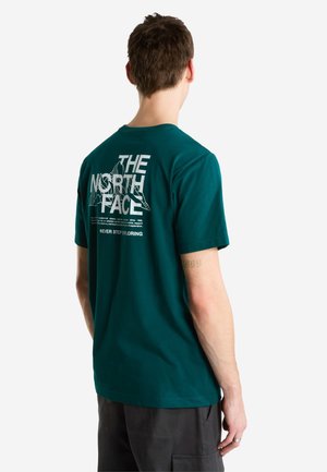 Hombre con una camiseta azul oscuro verdosa con el logo "THE NORTH FACE" y un gráfico de montaña impreso en blanco en la espalda, combinado con pantalones oscuros.