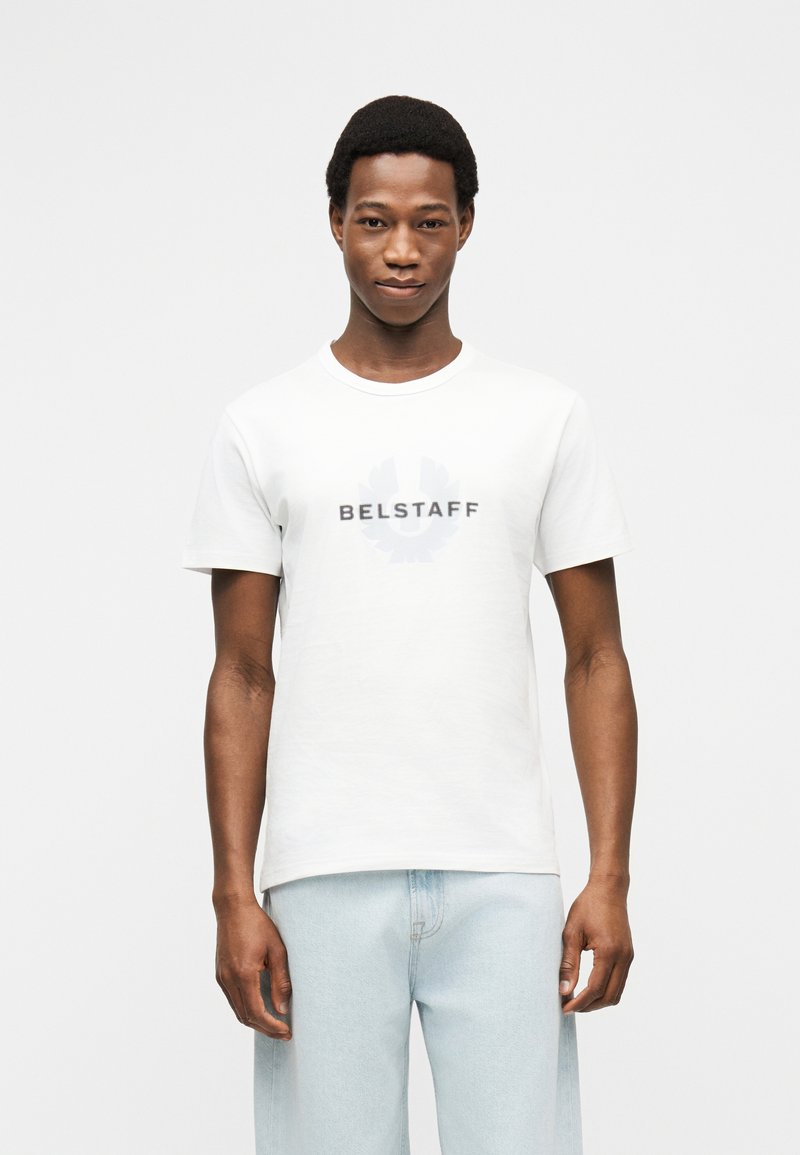 Νέος άνδρας που φοράει λευκό t-shirt με το λογότυπο Belstaff και ανοιχτόχρωμα τζιν, στέκεται μπροστά σε απλό, ανοιχτό φόντο.