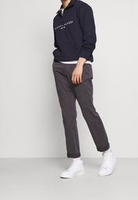 Felpa blu navy con zip e scritte bianche, dotata di una grande tasca a marsupio, abbinata a chinos grigi e sneakers bianche alte.