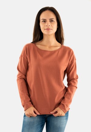 Femme aux longs cheveux noirs portant une chemise à manches longues couleur rouille et un jean bleu, se tenant contre un fond clair uni.