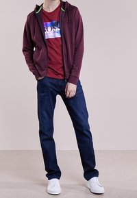 Sudadera burdeos con cremallera y acentos verdes sobre una camiseta gráfica roja, combinada con jeans azul oscuro y zapatillas blancas.