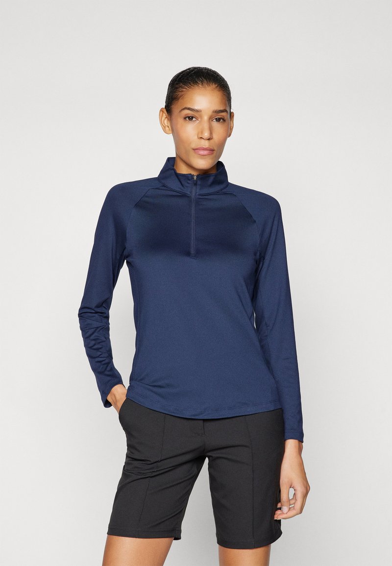 Tumedate pikkade varrukatega pluus, millel on tõmblukuga krae ja raglanvarrukad, koos mustade lühikeste pükstega. Kanga pind näib olema sile ja lähedane.