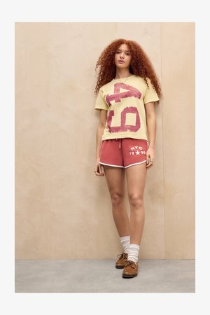 Ung kvinde med langt, krøllet rødt hår står op ad en trævæg iført en gul t-shirt med grafik, røde NYC-shorts, hvide sokker og brune sko.