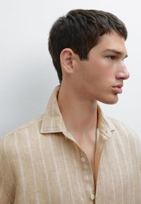 Beige linen shirt with white vertical stripes, button-up design, and a soft collar, featuring a subtle texture and relaxed fit.
