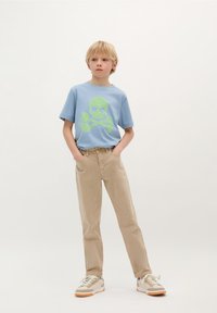 Blå t-shirt med kort ærme, der har et grønt dødningehoved og korslagte knogler grafik, parret med beige bukser og lyse sneakers.