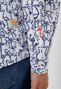 Camicia bianca con stampa di fenicotteri blu, con un'accenno di fenicottero arancione e un dettaglio rosso vicino al polsino, realizzata in tessuto leggero.