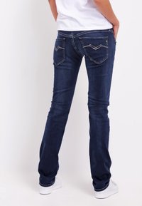 Vaqueros oscuros ajustados, con dos bolsillos traseros con costura decorativa, combinados con zapatillas blancas y una blusa clara.