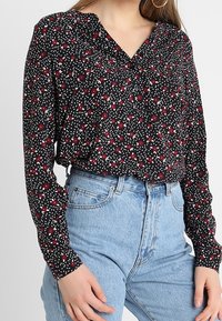 Blouse noire à manches longues avec un motif floral rouge, dotée d'un col en V, d'un tissu doux et d'une coupe ample, associée à un jean taille haute bleu clair.