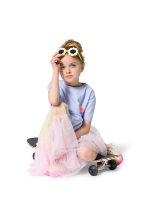 Παιδί καθισμένο σε σκέιτμπορντ, φοράει ανοιχτό μπλε πουκάμισο και πολύχρωμη φούστα από τούλι. Στρογγυλά γυαλιά ηλίου με διακοσμητικά λουλούδια ολο κληρώνουν την εμφάνιση.