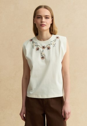 Frau trägt ärmelloses weißes Hemd mit Blumenstickerei am Ausschnitt und dunkelbraune Hose, steht vor beigem Hintergrund.