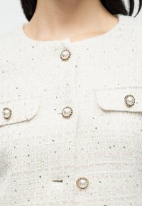 Veste blanche texturée avec des boutons effet perle, agrémentée d'accents dorés. Le design comprend deux poches avant avec des détails similaires aux boutons.