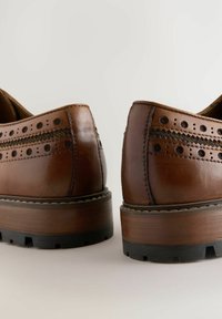 Chaussures en cuir marron avec des détails perforés, des coutures foncées et une épaisse semelle en caoutchouc noir. Forme arrondie avec un talon bas.