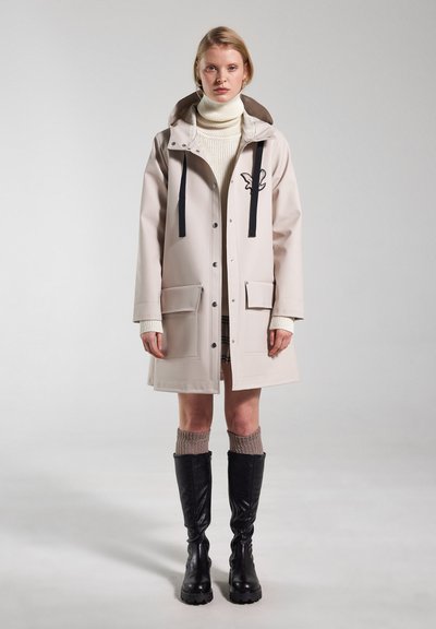 Beige regenjas met zwarte accenten, twee voorzakken, drukknoopsluitingen en een capuchon. Draag boven een witte coltrui, gecombineerd met hooggeschoeide zwarte laarzen.