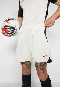 Hvite Nike-shorts med svarte sideakenter, en elastisk livlinje og en rød logo. Svarte sokker og en mønstrete fotball i hånden.