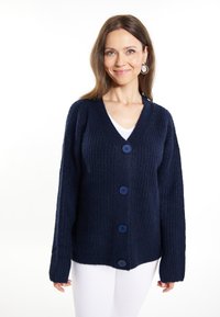 Granatowy sweter z dzianiny z dekoltem w serek, długimi rękawami i pięcioma niebieskimi guzikami. Struktura prążkowana i luźny krój. Połączony z białą bluzką i spodniami.