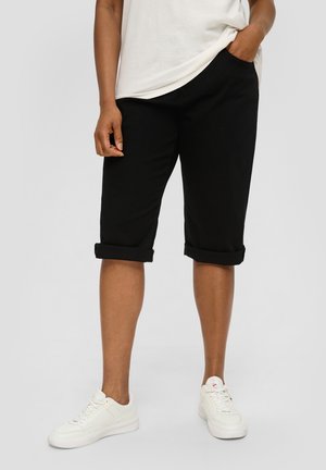 Personne debout portant un short bermuda noir roulé, des baskets blanches et une chemise blanche avec une main dans la poche, sur un fond uni.