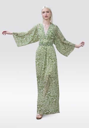 Mujer con cabello largo y rubio, vestida con un vestido largo de manga ancha con un patrón verde y blanco, de pie con los brazos extendidos hacia los lados.