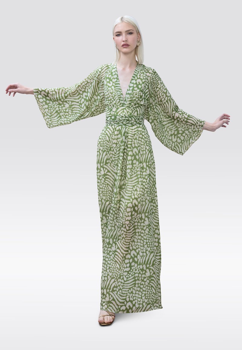 Žena s dlhými blond vlasmi, oblečená v zelenej a bielej vzorovanej dlhej sukni s širokými rukávmi, stojaca s natiahnutými rukami do strán.