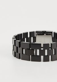 Svart armbåndsur med rektangulært design, med glatte, polerte lenker og en minimalistisk lås. Sølvfarget bakside med merkegraveringer.