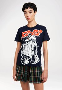 Woman with short black hair wearing a navy shirt printed with R2-D2 and a green plaid short skirt, standing against a white background.