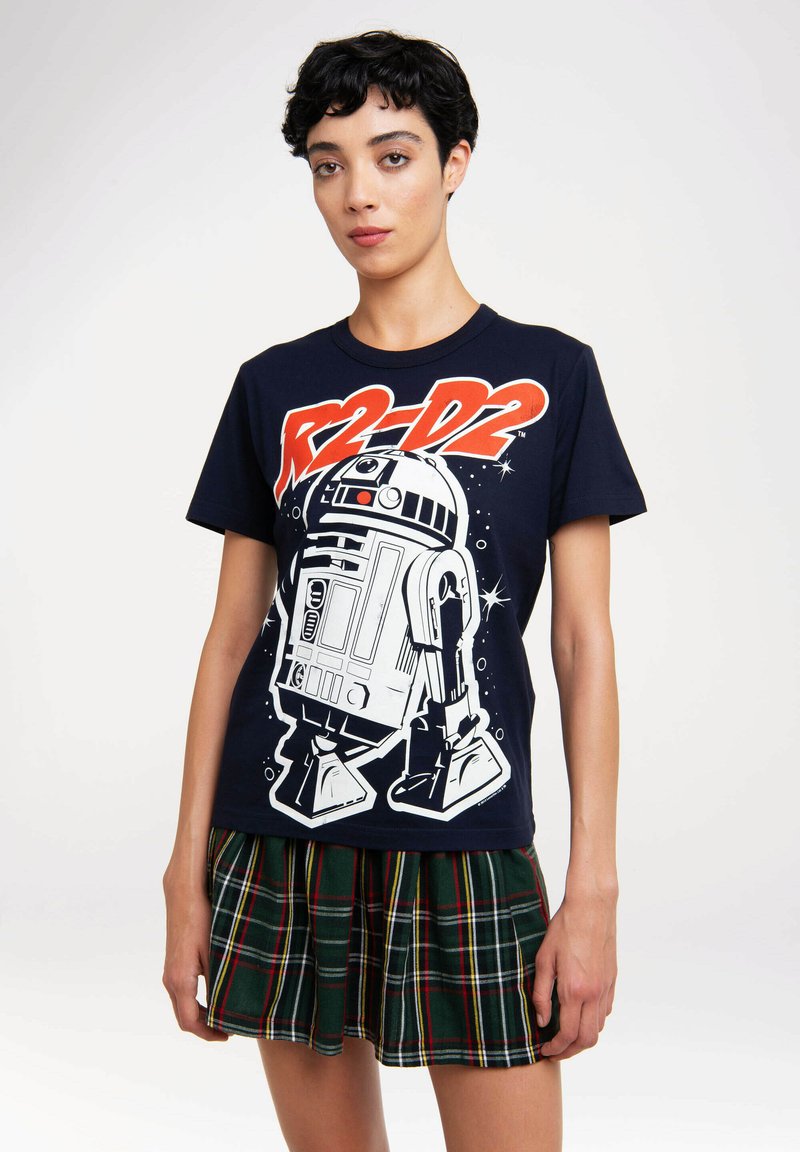 Woman with short black hair wearing a navy shirt printed with R2-D2 and a green plaid short skirt, standing against a white background.