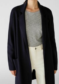Marinblå stickad cardigan med öppen front och stora slag, lagd över en randig skjorta. Vita höga jeans fulländar outfiten.