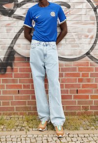 Jersey azul de Nike con detalles en blanco y dorado, vaqueros holgados de lavado claro y zapatillas marrones y azul turquesa, de pie contra una pared de ladrillo.