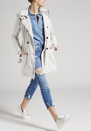 Lichtgewicht, beige trenchcoat met knoopdetails, gecombineerd met een denim overhemd, versleten blauwe jeans en witte sneakers.