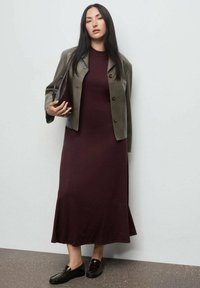 Femme aux longs cheveux noirs portant une robe bourgogne, une veste courte olive, des mocassins noirs et tenant un sac à main marron foncé.