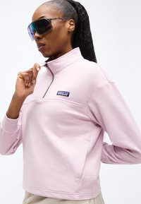 Γυναίκα με μεγάλα ανακλαστικά γυαλιά ηλίου και ανοιχτό ροζ πουλόβερ Patagonia με φερμουάρ στο λαιμό, που ρυθμίζει το φερμουάρ με το ένα χέρι.