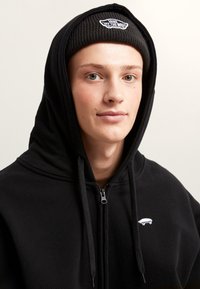 Felpa nera con zip e tasca a marsupio, con una piccola grafica bianca di uno skateboard. Indossata con un berretto a costine nero.