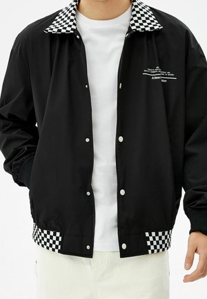 Schwarze Jacke mit Knöpfen, schwarzem und weiß kariertem Kragen und Saum, getragen über einem weißen T-Shirt, zeigt den Oberkörper.