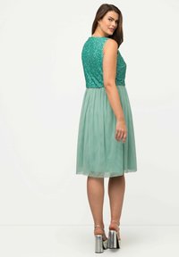 Vestido de cuerpo de lentejuelas verdes con una falda de gasa menta fluida. Presenta un escote redondeado en la parte delantera y trasera, combinado con sandalias de tacón cuadrado plateadas.