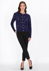Tengerészkék kardigán kerek nyakkivágással, gyöngygombokkal és díszes fehér virágmintákkal. Fekete, szabott nadrággal és magassarkú cipővel párosítva.