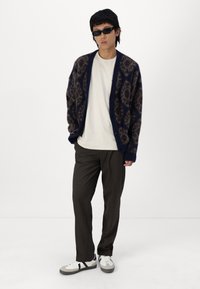 Navy patterned cardigan with brown motifs, worn over a cream t-shirt. Paired with dark trousers and white sneakers with gray accents.