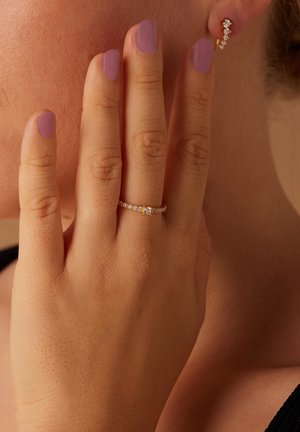 Gouden ring met kleine heldere stenen, gedragen aan een hand met lavendelkleurige nagellak, gedeeltelijk bedekt door vingers op een neutrale achtergrond.