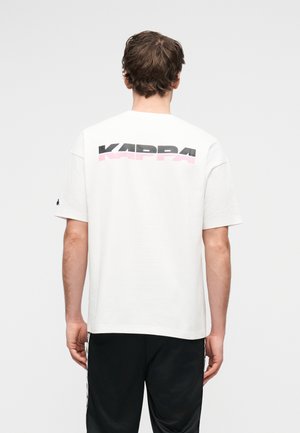 Άνδρας που φορά λευκό μπλουζάκι με μαύρη και ροζ επιγραφή "KAPPA" στην πλάτη και μαύρο παντελόνι με ρίγες στο πλάι, με την πλάτη γυρισμένη, μπροστά σε απλό φόντο.