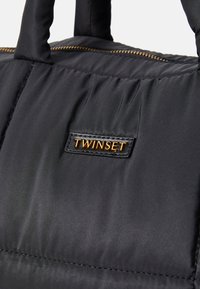 Close-up of a black padded bag with quilted texture, gold zipper, and a leather patch label with gold letters reading "TWINSET."